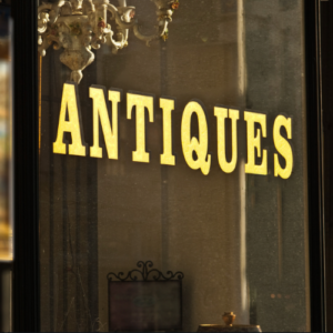 Close-up of gold "Antiques" lettering on a shop window, with a glimpse of a chandelier and other objects inside.