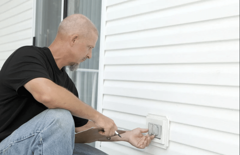 A man repairing or inspecting an exterior electrical outlet on a white house wall.