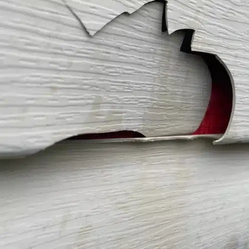 Cracked and warped white vinyl siding with a large hole exposing red underlayment, requiring emergency siding repair Shawnee