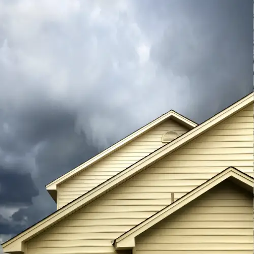 Dramatic view of a home's roofline and tan siding set against dark, heavy storm clouds, symbolizing the need for proactive roofing storm damage repair by Xtreme Exteriors in Nixa, MO