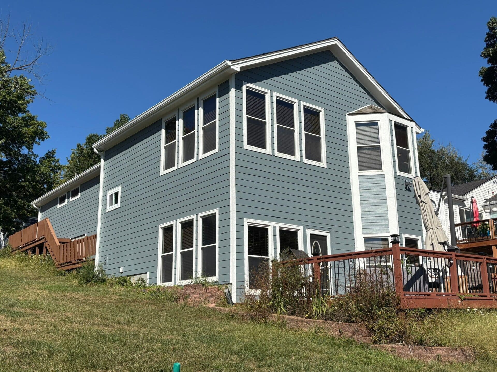 Back corner of a two-story home after a full James Hardie reside, showing new blue exterior boards, white trim, updated windows, and a brown elevated deck, photographed on a sunny day