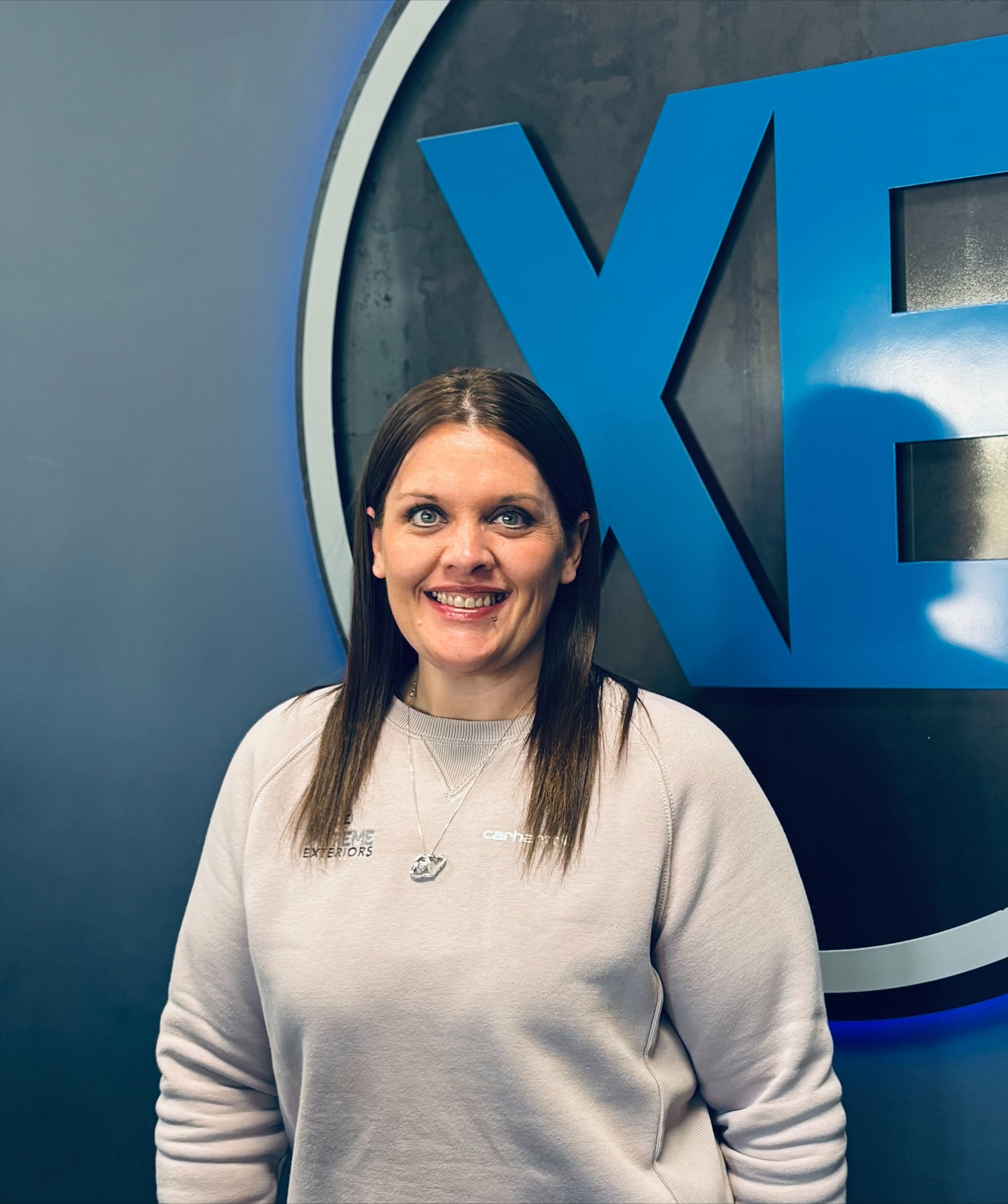 Xtreme Exteriors Shawnee Branch Manager, Dani Shelton, is smiling and standing in front of a blue XE wearing a gray carhartt sweatshirt with Xtreme Exteriors embroidered on the right left chest